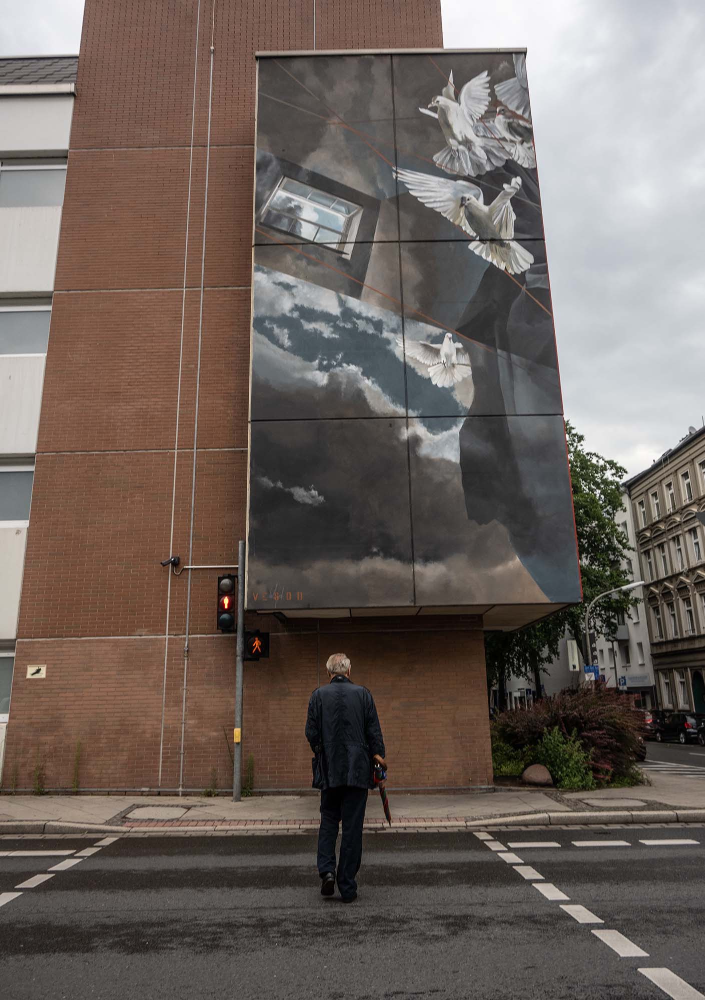 Die Katze spaziert über eine Straße. Über ihm ein Mural von weißen Tauben im Flug und einem Fenster.