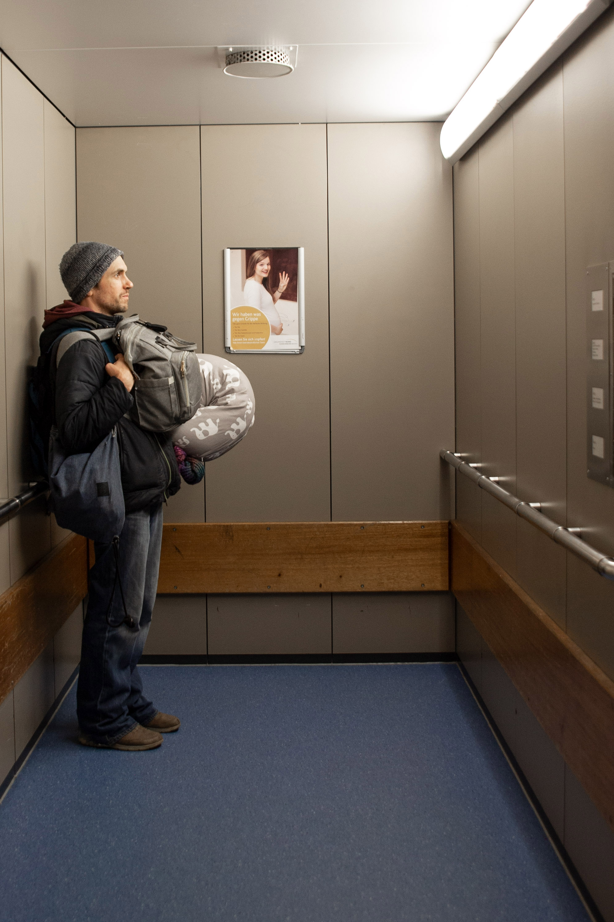 Vollgepackter Ehemann im Aufzug. Eine schwangere Frau winkt von einem Plakat in die Kamera