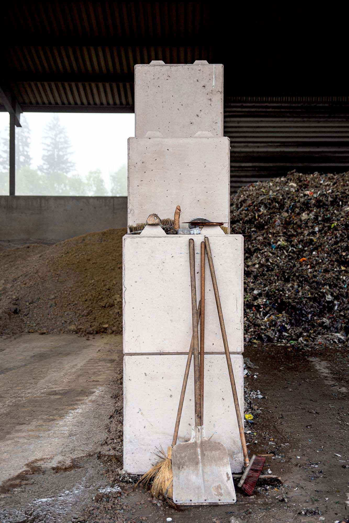 Eine Trennwand einer Lagerhalle eines Wertstoffhofes, an welcher zwei Besen und eine Schaufel lehnen. Auf einem Absatz stehen weitere Dinge. Auch ein Dildo.